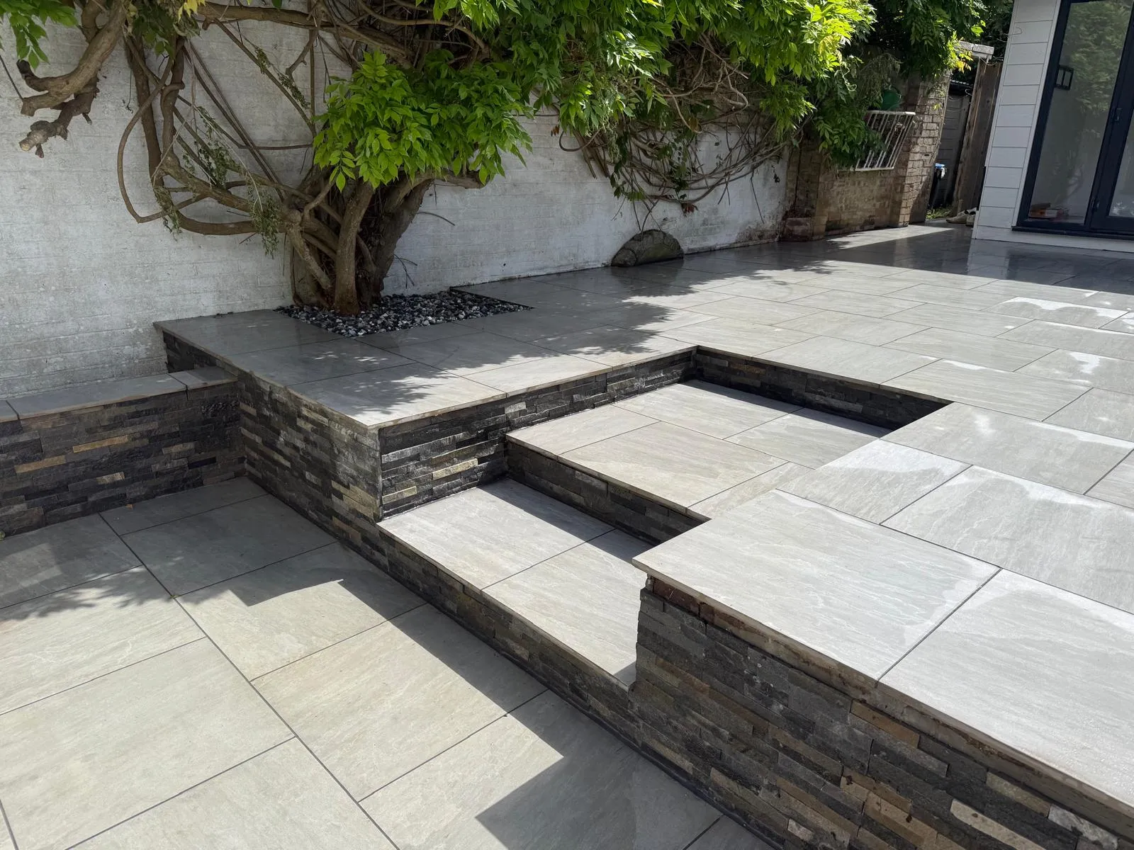 Split-face stone-clad raised bed around a mature wisteria on a grey porcelain patio in Farnborough Village