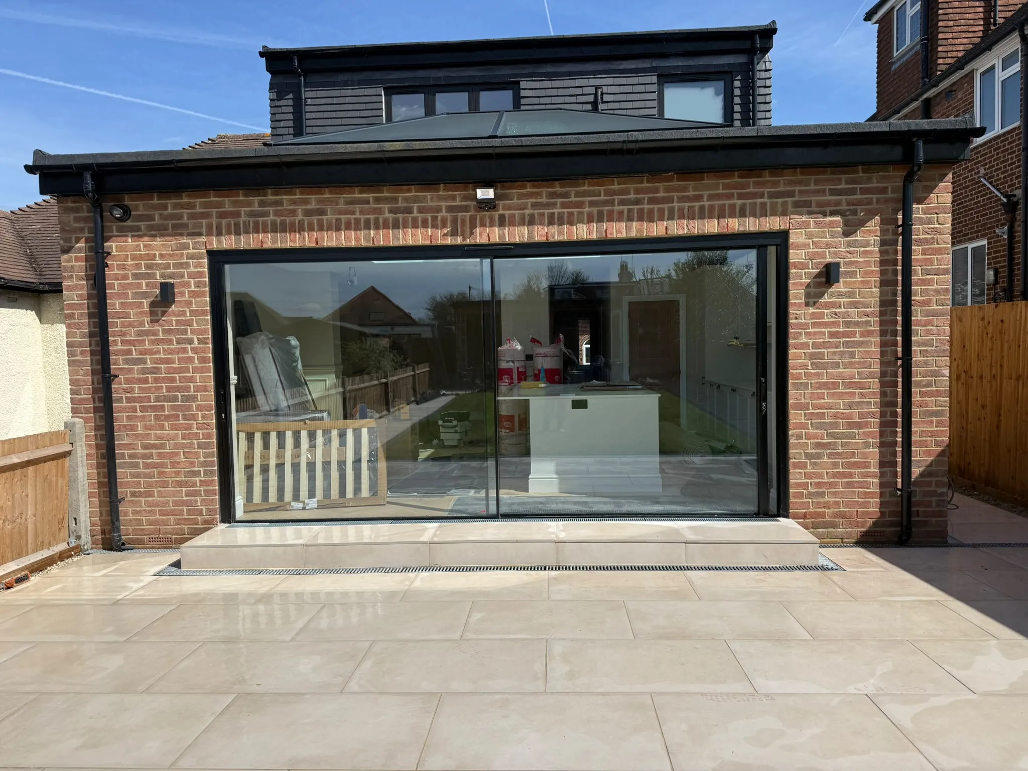 Cream porcelain patio and brick garden-room extension under blue sky by Prestige Landscapes
